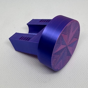 May include: A purple bottle stopper with a starburst design. The stopper has a cylindrical body and a rectangular base with a series of parallel lines. The starburst design is in a lighter shade of purple.
