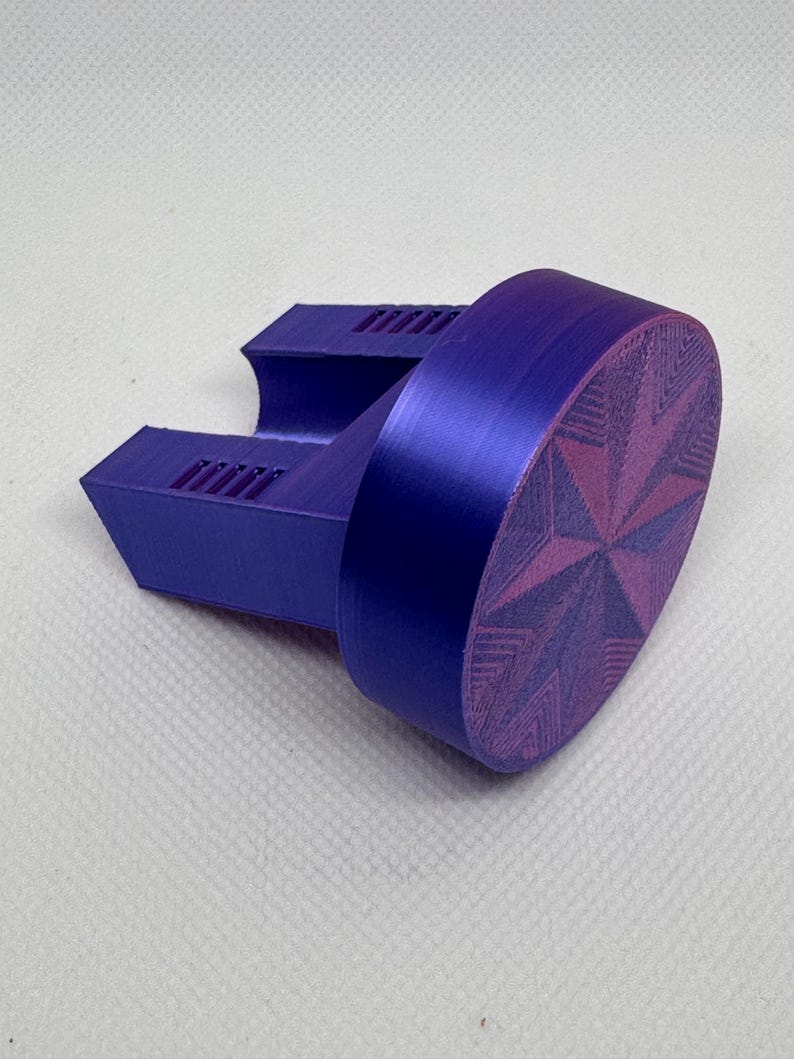 May include: A purple bottle stopper with a starburst design. The stopper has a cylindrical body and a rectangular base with a series of parallel lines. The starburst design is in a lighter shade of purple.