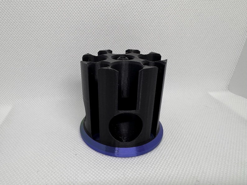 Puede incluir: Un objeto cil&iacute;ndrico negro con una serie de ranuras verticales y una base circular con un borde azul. La parte superior tiene un patr&oacute;n en forma de estrella. El objeto parece estar impreso en 3D.