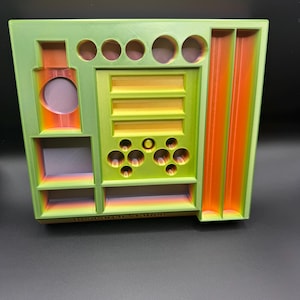 Puede incluir: Un organizador verde y naranja impreso en 3D con varios compartimentos, que incluyen agujeros circulares, estantes y ranuras verticales. El texto "Research Station" es visible en la parte inferior. El organizador est&aacute; dise&ntilde;ado para guardar objetos peque&ntilde;os.
