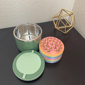 May include: A green and silver ashtray set with a matching lid and a multi-colored, round container. A gold geometric sculpture sits nearby. The ashtray set is on a dark surface.