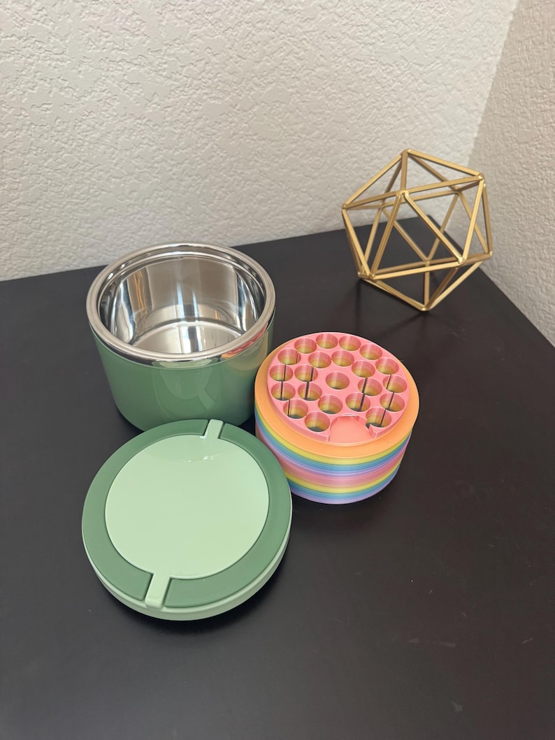May include: A green and silver ashtray set with a matching lid and a multi-colored, round container. A gold geometric sculpture sits nearby. The ashtray set is on a dark surface.