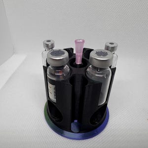 Puede incluir: Un soporte de viales negro impreso en 3D con una base circular y seis compartimentos. Cuatro viales de vidrio con tapas plateadas est&aacute;n en el soporte, junto con una pipeta rosa. La base tiene un degradado de azul, verde y morado.