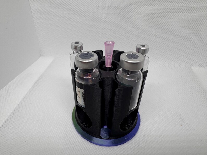 Puede incluir: Un soporte de viales negro impreso en 3D con una base circular y seis compartimentos. Cuatro viales de vidrio con tapas plateadas est&aacute;n en el soporte, junto con una pipeta rosa. La base tiene un degradado de azul, verde y morado.