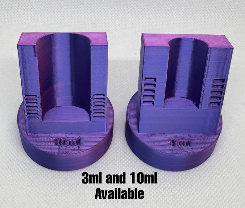May include: Two purple 3D-printed measuring tools. One is labeled "10 ml" and the other "3 ml". The text "3ml and 10ml Available" is printed below the tools. Each tool has a cylindrical opening and a rectangular body with vertical slots.