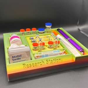 Puede incluir: Un organizador de estaci&oacute;n de investigaci&oacute;n impreso en 3D en colores verde, amarillo y arco&iacute;ris. Contiene viales, jeringas, toallitas con alcohol y un bol&iacute;grafo morado. El texto "Research Station" es visible en la parte delantera.