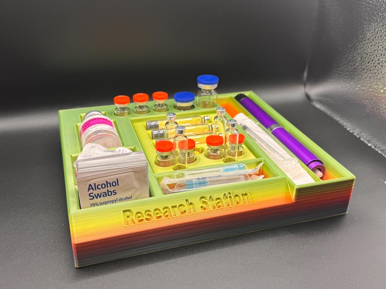 Puede incluir: Un organizador de estaci&oacute;n de investigaci&oacute;n impreso en 3D en colores verde, amarillo y arco&iacute;ris. Contiene viales, jeringas, toallitas con alcohol y un bol&iacute;grafo morado. El texto "Research Station" es visible en la parte delantera.
