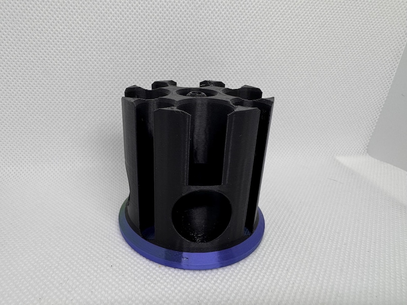Puede incluir: Un objeto cil&iacute;ndrico negro con una base azul. La parte superior tiene m&uacute;ltiples aberturas y los lados ranuras verticales. El objeto parece estar impreso en 3D y probablemente sea un soporte o organizador.