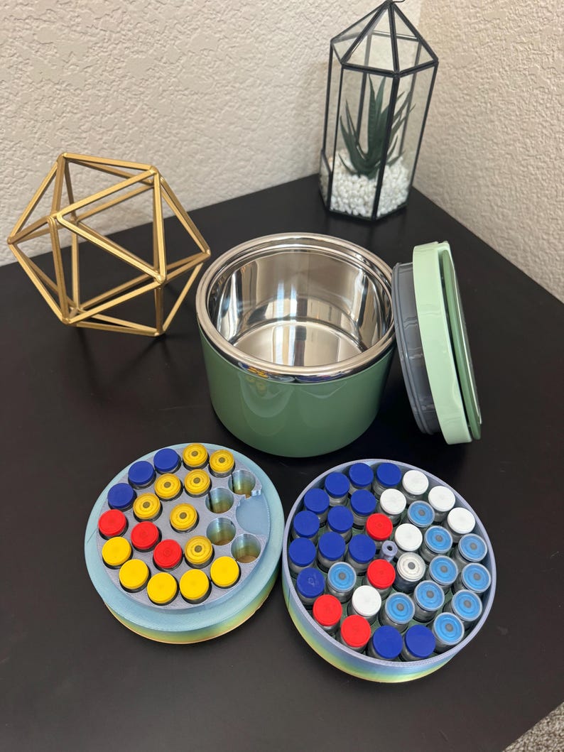 May include: An open, green insulated food container with a stainless steel interior sits next to two circular containers filled with small vials. The vials have colorful caps in red, yellow, blue, and white.