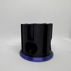 Puede incluir: Un objeto cil&iacute;ndrico negro con una serie de recortes y una base circular con un borde p&uacute;rpura. El objeto tiene un dise&ntilde;o geom&eacute;trico y parece estar impreso en 3D. El fondo es una superficie blanca lisa.