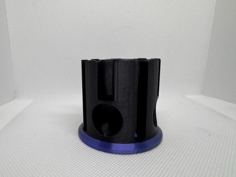 Puede incluir: Un objeto cil&iacute;ndrico negro con una serie de recortes y una base circular con un borde p&uacute;rpura. El objeto tiene un dise&ntilde;o geom&eacute;trico y parece estar impreso en 3D. El fondo es una superficie blanca lisa.