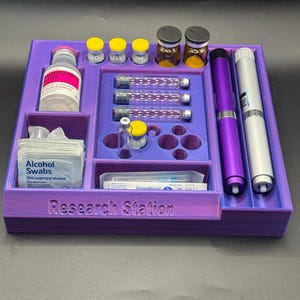 Puede incluir: Un organizador morado impreso en 3D, llamado "Research Station", que contiene viales, jeringas, toallitas con alcohol y dos bol&iacute;grafos. El organizador tiene m&uacute;ltiples compartimentos para diversos suministros m&eacute;dicos o de investigaci&oacute;n.