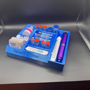 Puede incluir: Un organizador azul de estaci&oacute;n de investigaci&oacute;n impreso en 3D con compartimentos que contienen viales, jeringas, toallitas con alcohol y un bol&iacute;grafo morado. El texto "Research Station" est&aacute; impreso en la parte delantera.