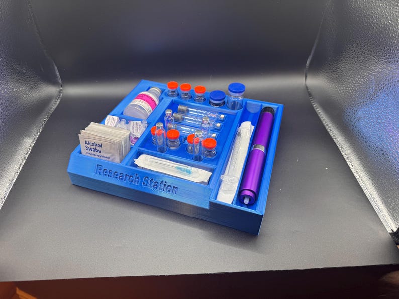 Puede incluir: Un organizador azul de estaci&oacute;n de investigaci&oacute;n impreso en 3D con compartimentos que contienen viales, jeringas, toallitas con alcohol y un bol&iacute;grafo morado. El texto "Research Station" est&aacute; impreso en la parte delantera.