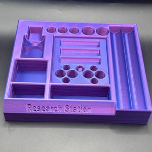 Puede incluir: Un organizador de Research Station impreso en 3D en morado y azul. El organizador tiene varios compartimentos, incluyendo agujeros circulares, ranuras rectangulares y recortes de botones estilo controlador. El texto "Research Station" est&aacute; impreso en la parte delantera.
