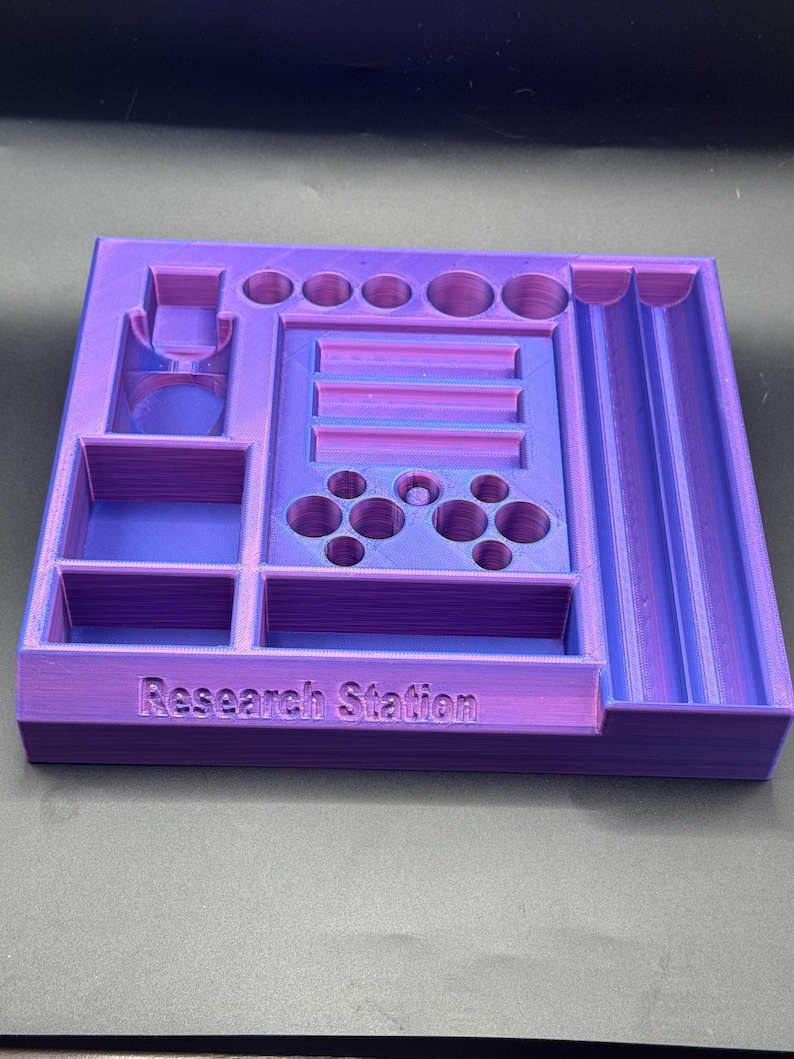 Puede incluir: Un organizador de Research Station impreso en 3D en morado y azul. El organizador tiene varios compartimentos, incluyendo agujeros circulares, ranuras rectangulares y recortes de botones estilo controlador. El texto "Research Station" est&aacute; impreso en la parte delantera.