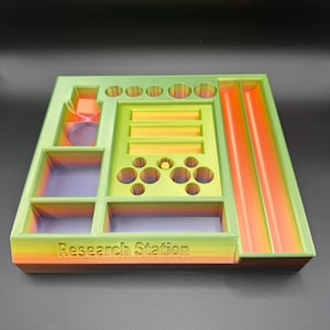 Puede incluir: Un organizador multicolor impreso en 3D con el texto "Research Station". El organizador presenta varios compartimentos, incluyendo agujeros circulares, ranuras rectangulares y un canal inclinado. Los colores transicionan de naranja a verde, con acentos morados y amarillos.