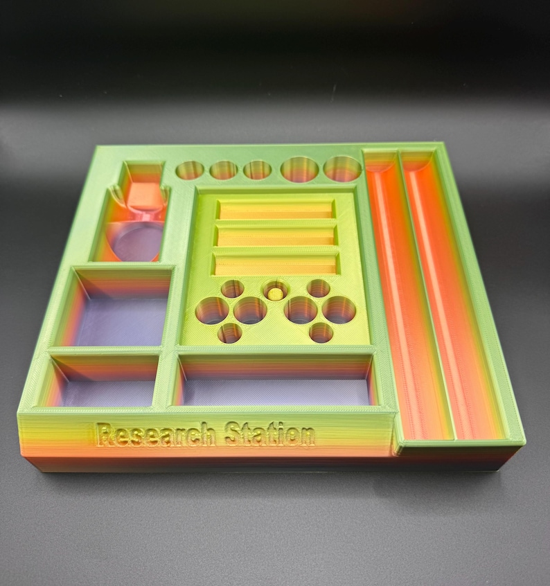 Puede incluir: Un organizador multicolor impreso en 3D con el texto "Research Station". El organizador presenta varios compartimentos, incluyendo agujeros circulares, ranuras rectangulares y un canal inclinado. Los colores transicionan de naranja a verde, con acentos morados y amarillos.