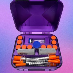 Op de afbeelding: Een paarse medische reiskoffer, open om georganiseerde compartimenten te laten zien. Binnenin zitten oranje dopjes, spuiten, alcoholdoekjes en een kleine glazen fles met een blauwe dop. De koffer heeft zwarte sluitingen.