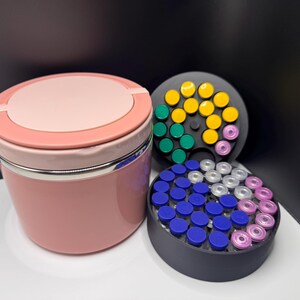 May include: A pink and silver container with a matching lid, next to two black circular holders filled with colorful vials. The vials have blue, yellow, green, white, and pink caps. The items are on a white surface.