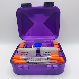 May include: A purple hard-shell storage case containing medical supplies. Inside are two stacks of white packets labeled Alcohol Swabs, several orange-capped syringes, small orange-capped vials, and a central vial with a blue cap.