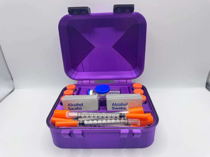 May include: A purple hard-shell storage case containing medical supplies. Inside are two stacks of white packets labeled Alcohol Swabs, several orange-capped syringes, small orange-capped vials, and a central vial with a blue cap.