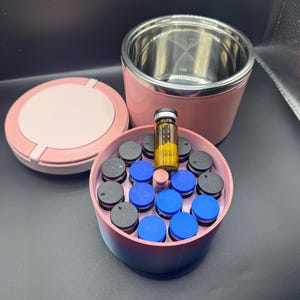 May include: A pink and stainless steel insulated container with a matching lid. Inside, a circular container holds small vials with blue and gray caps, and one vial with a yellow liquid. A single vial with a yellow liquid sits on top.