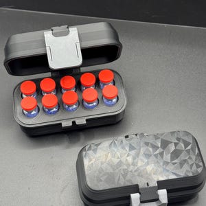 Peut inclure: Un étui de rangement rectangulaire noir avec un couvercle à charnière et un loquet, contenant dix petits flacons en verre avec des bouchons rouge vif. Un deuxième étui identique est fermé, présentant un motif géométrique texturé.