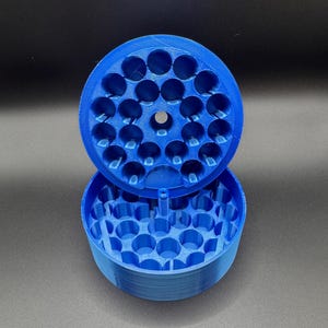 May include: A blue 3D-printed container with a lid, featuring multiple circular compartments. The lid is open, revealing the interior. The container is designed to hold small items, with a cylindrical shape.
