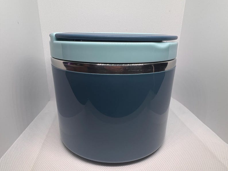 May include: A cylindrical, insulated food container in shades of blue and teal. The container has a stainless steel band and a matching lid. The design is sleek and modern, suitable for carrying lunches or snacks.