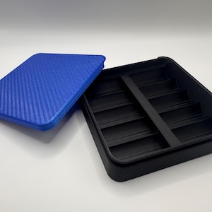 May include: A black rectangular storage container with a lid. The lid is blue with a textured, woven pattern. The container has several vertical dividers inside. The lid is slightly ajar, revealing the interior.