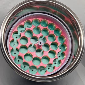 May include: A stainless steel tumbler with a pink and green insert. The insert has a circular design with multiple green, rounded protrusions. The tumbler is set against a light gray background.