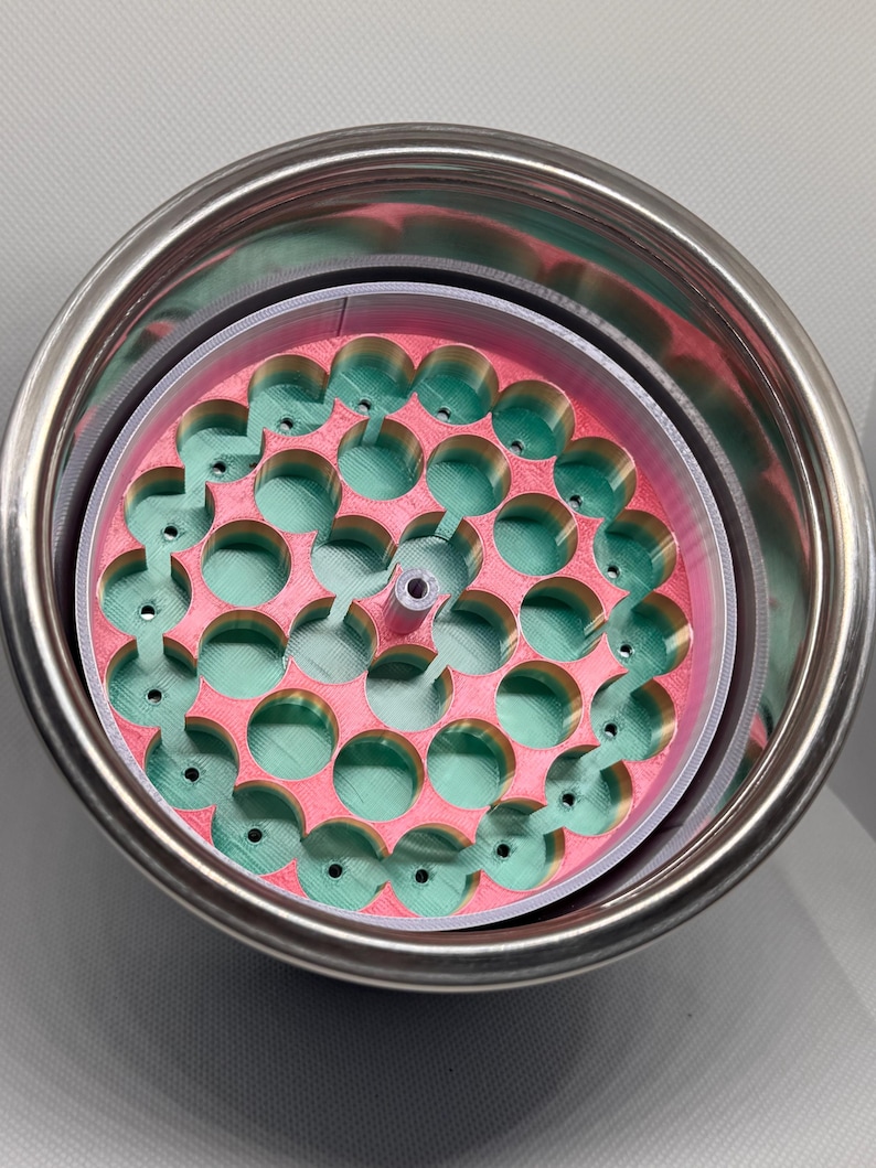 May include: A stainless steel tumbler with a pink and green insert. The insert has a circular design with multiple green, rounded protrusions. The tumbler is set against a light gray background.