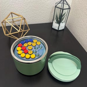 May include: A green container with a lid, filled with colorful, round objects. The container is open, revealing a blue interior with yellow, red, and blue items. A gold geometric shape and a terrarium with a plant are in the background.