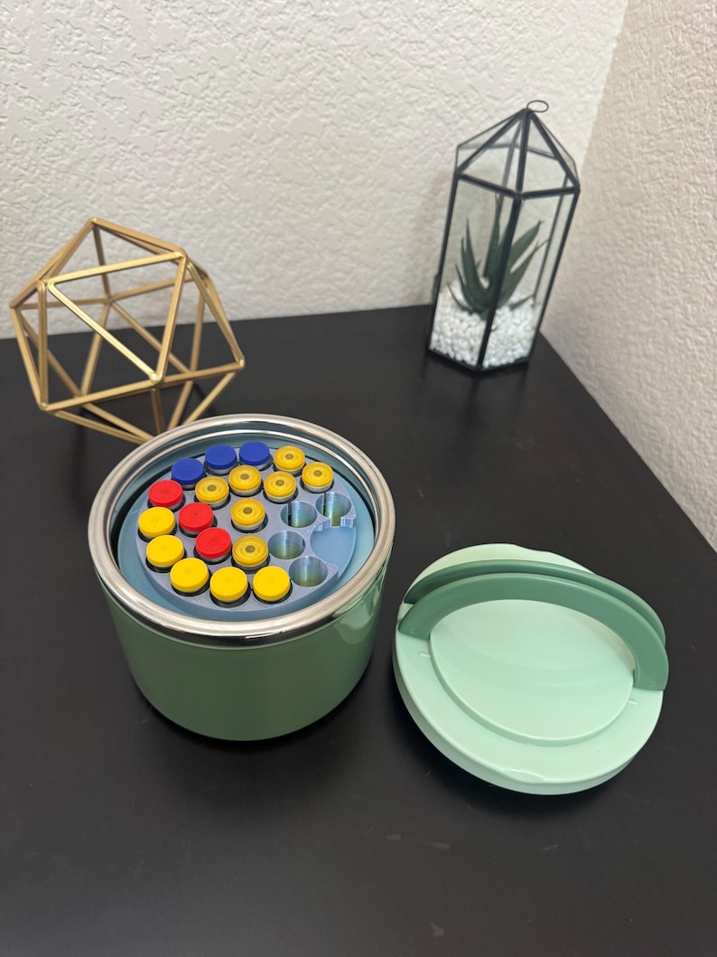 May include: A green container with a lid, filled with colorful, round objects. The container is open, revealing a blue interior with yellow, red, and blue items. A gold geometric shape and a terrarium with a plant are in the background.