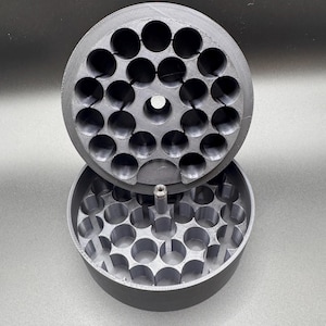 May include: A gray, 3D-printed container with a lid. The container and lid are round and feature multiple circular holes. The lid is partially open, revealing the interior structure. The design suggests a storage or organizational function.