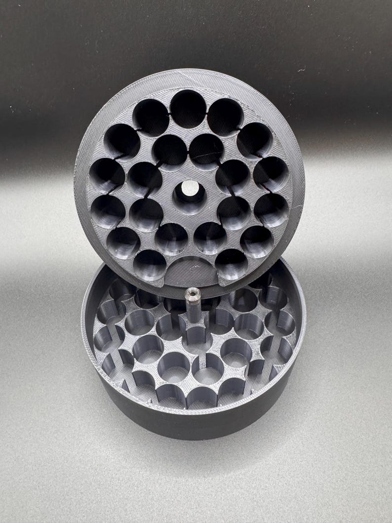 May include: A gray, 3D-printed container with a lid. The container and lid are round and feature multiple circular holes. The lid is partially open, revealing the interior structure. The design suggests a storage or organizational function.