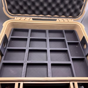 Puede incluir: Maleta rígida beige abierta con un inserto gris oscuro con múltiples compartimentos rectangulares. La maleta tiene un forro de espuma negra en la tapa. La maleta está diseñada para el almacenamiento y la organización.