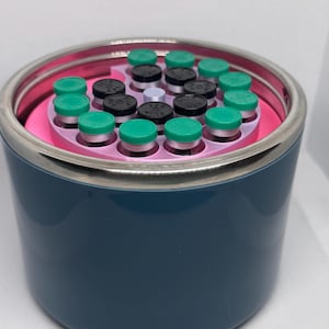 May include: A dark blue cylindrical container with a silver rim, holding multiple small vials. The vials have green and black caps, arranged in a pink circular holder. The container is on a white surface.