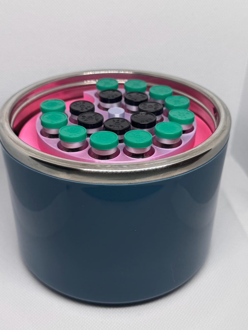 May include: A dark blue cylindrical container with a silver rim, holding multiple small vials. The vials have green and black caps, arranged in a pink circular holder. The container is on a white surface.
