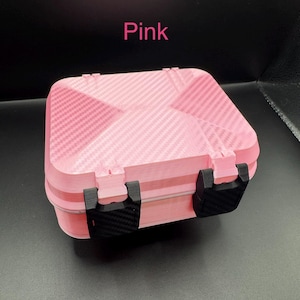 May include: A rectangular pink storage case with a carbon fiber texture on the lid and base. The case features two black latches on the front and the word Pink printed in pink text above the object. The design has a geometric pattern.