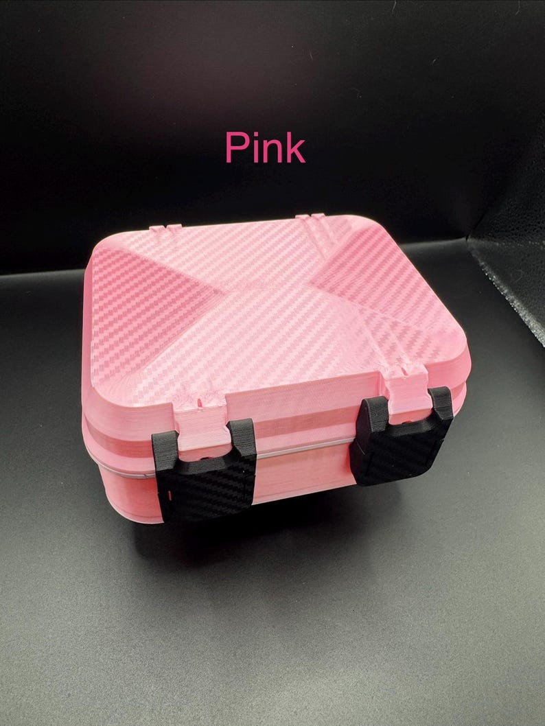 May include: A rectangular pink storage case with a carbon fiber texture on the lid and base. The case features two black latches on the front and the word Pink printed in pink text above the object. The design has a geometric pattern.