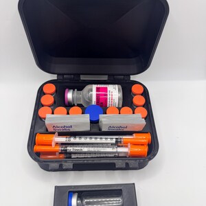May include: A black carrying case containing medical supplies including a glass vial with a pink label, multiple small orange-capped vials, two stacks of alcohol swabs, and three Easy-Touch syringes with orange caps. A separate black holder holds one blue-capped vial.