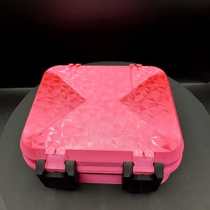 May include: A square, bright pink storage case featuring a geometric, faceted, low-poly texture on the lid. The container is secured with two black, rectangular latches positioned on the front edge, providing a contrast to the vibrant pink color.
