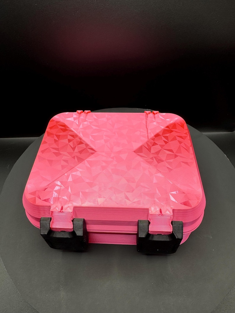 May include: A square, bright pink storage case featuring a geometric, faceted, low-poly texture on the lid. The container is secured with two black, rectangular latches positioned on the front edge, providing a contrast to the vibrant pink color.