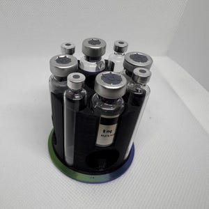 Puede incluir: Un organizador circular negro que contiene varios viales de vidrio peque&ntilde;os con tapas plateadas. El organizador tiene un borde verde y azul. Un vial tiene una etiqueta con texto. Los viales est&aacute;n dispuestos en un patr&oacute;n circular.