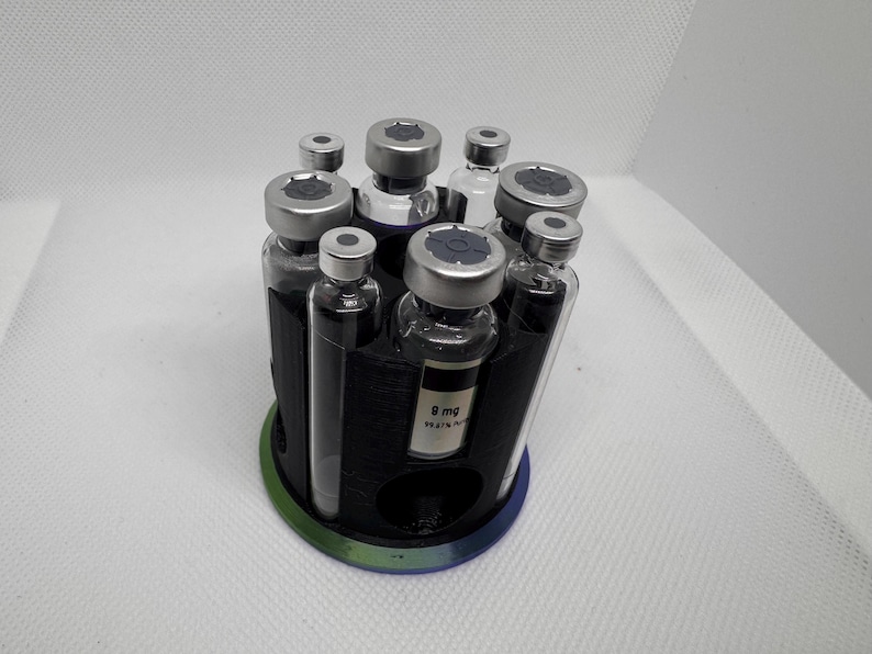 Puede incluir: Un organizador circular negro que contiene varios viales de vidrio peque&ntilde;os con tapas plateadas. El organizador tiene un borde verde y azul. Un vial tiene una etiqueta con texto. Los viales est&aacute;n dispuestos en un patr&oacute;n circular.
