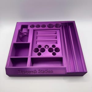 Puede incluir: Un organizador de Research Station morado impreso en 3D con varios compartimentos, incluyendo soportes circulares, una ranura y una secci&oacute;n con m&uacute;ltiples agujeros. El texto "Research Station" est&aacute; impreso en la parte delantera.