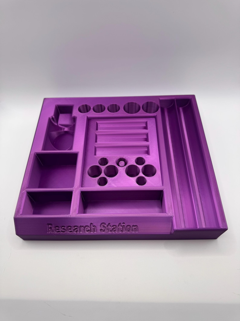 Puede incluir: Un organizador de Research Station morado impreso en 3D con varios compartimentos, incluyendo soportes circulares, una ranura y una secci&oacute;n con m&uacute;ltiples agujeros. El texto "Research Station" est&aacute; impreso en la parte delantera.