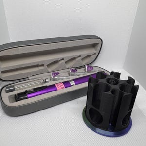 Puede incluir: Un bol&iacute;grafo m&eacute;dico morado y accesorios se muestran en un estuche gris abierto. A la derecha hay un soporte cil&iacute;ndrico negro con m&uacute;ltiples aberturas. El estuche contiene un bol&iacute;grafo, viales y otros suministros m&eacute;dicos.